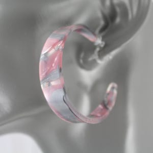 May include: A close-up of a pink, gray, and clear hoop earring. The earring is a semi-circle shape with a marbled design. The earring is attached to a blurred ear in the background.