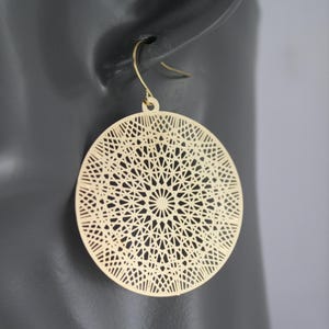 May include: Gold-colored, circular earrings with an intricate, geometric openwork design. The earrings have a hook closure and are displayed against a dark gray background. The earrings are a stylish accessory.