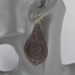 May include: A pair of brown dangle earrings with a delicate, intricate design. The earrings are made of metal and have a teardrop shape. The design features a circular pattern with a starburst in the center.