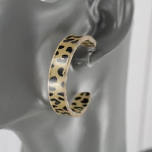 May include: A pair of hoop earrings with a leopard print design. The earrings are a light tan color with black spots. The earrings are shown on a gray mannequin head.