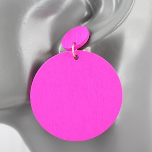 May include: Bright pink wooden earrings. Each earring features a small, round stud and a larger, circular pendant. The earrings are attached with a gold-colored hook. The earrings are a vibrant accessory.
