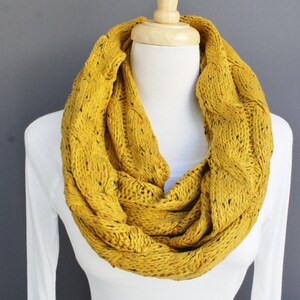 May include: A yellow knitted infinity scarf with a cable knit pattern and black and red flecks.
