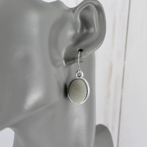 May include: A single silver-tone earring with a small, oval, light green stone. The stone is set within a silver frame and hangs from a simple hook. The earring is displayed on a gray mannequin head.