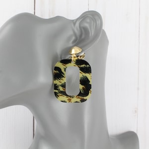 May include: A pair of gold clip-on earrings with a square-shaped design. The earrings are decorated with a leopard print pattern in black, brown, and yellow.