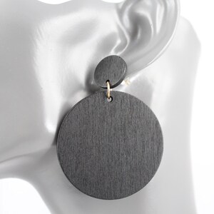 May include: Dark gray wooden earrings with a circular design. The earrings feature a small, round stud connected to a larger, circular pendant. The earrings are suspended from a gold-colored hook. The wood grain is visible on the surface.