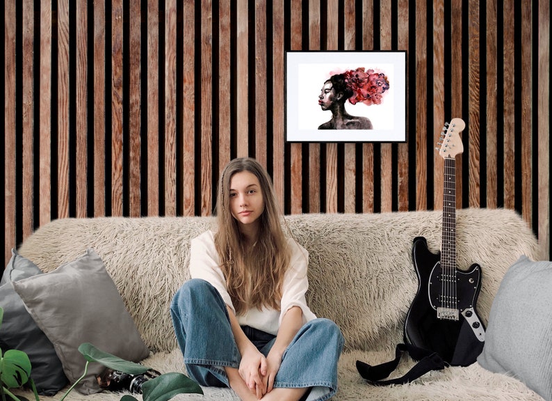May include: A woman sits on a couch with a guitar and a framed watercolor painting of a woman with red hair. The painting is on a wood panel wall.