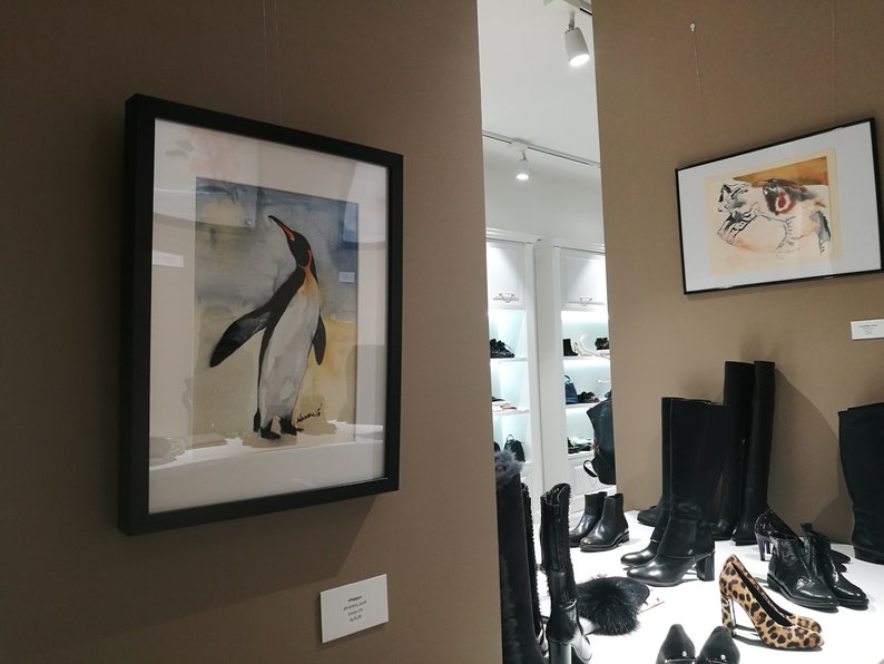 Puede incluir: Una pintura de acuarela enmarcada de un ping&uuml;ino de pie en una playa de arena. La pintura est&aacute; colgada en una pared marr&oacute;n en una tienda con botas y zapatos negros en exhibici&oacute;n.