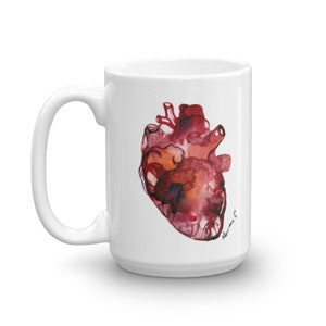 以下が含まれることがあります： 赤い人間の心臓の水彩画が描かれた白いセラミックマグカップ。心臓は赤、オレンジ、茶色の色合いで描かれています。