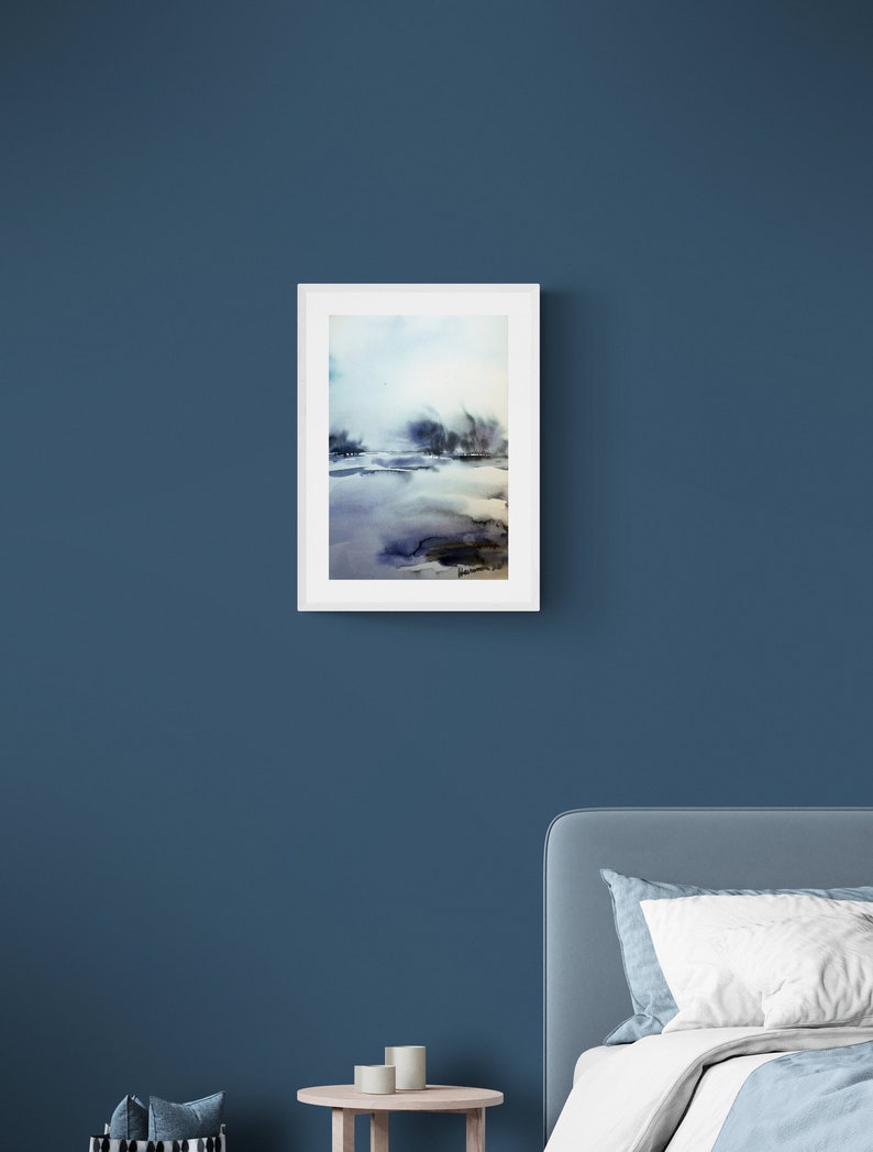 May include: A framed watercolour painting depicting a landscape. The artwork uses blues, purples, and browns to create a misty, atmospheric effect. The white frame contrasts with the dark blue wall, creating a striking visual.