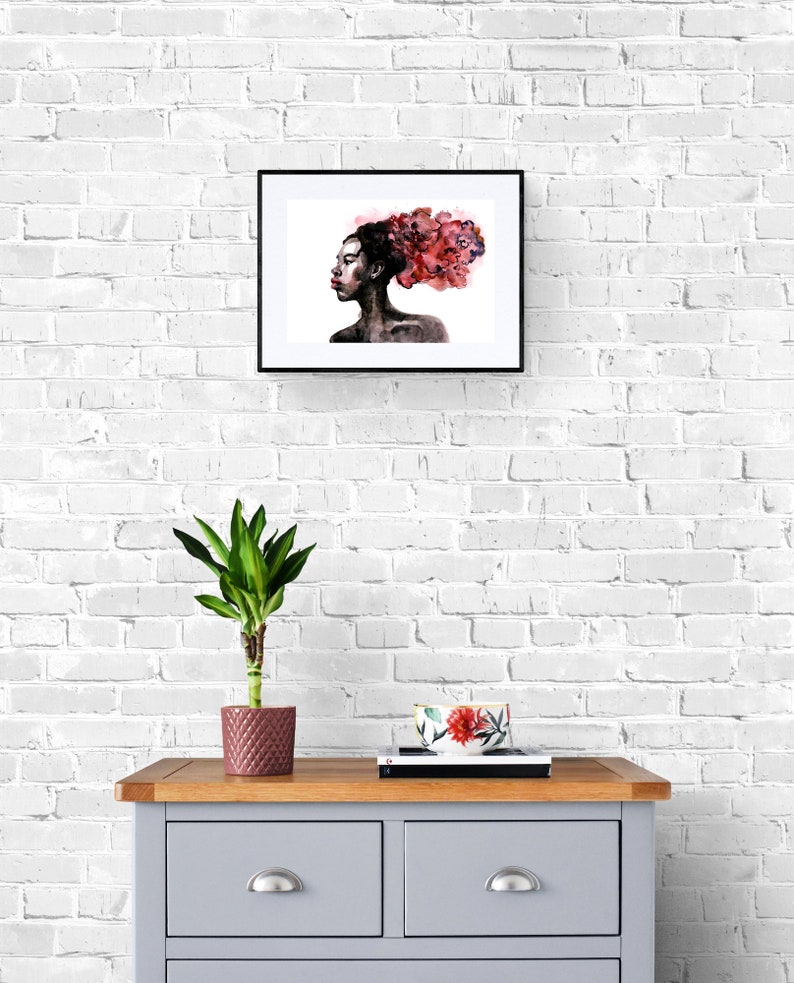 May include: A framed watercolor painting of a woman with a pink and red floral design in her hair. The painting is hanging on a white brick wall above a gray dresser with a plant in a pink pot and a floral bowl on top.