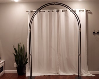 May include: A black metal archway with two white curtains hanging from it. The archway is standing on a wooden floor with a rug in front of it.