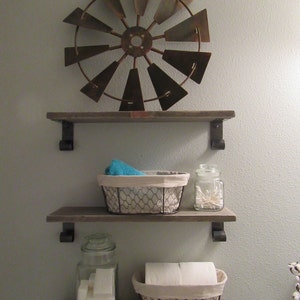 May include: Three wooden shelves with metal brackets, mounted on a light grey wall. The top shelf has a rustic metal windmill decoration. The middle shelf has a wire basket with a blue towel and a glass jar with white cotton buds. The bottom shelf has a glass jar with white soap bars and a wire basket with a white roll of toilet paper.