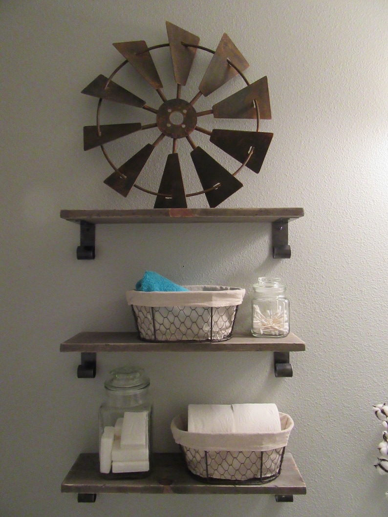 May include: Three wooden shelves with metal brackets, mounted on a light grey wall. The top shelf has a rustic metal windmill decoration. The middle shelf has a wire basket with a blue towel and a glass jar with white cotton buds. The bottom shelf has a glass jar with white soap bars and a wire basket with a white roll of toilet paper.