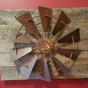 May include: A rustic metal windmill wall decor with eight blades, mounted on a weathered wood background. The blades are a dark brown colour with a hint of rust. The wood background is a light brown colour with a grey patina.