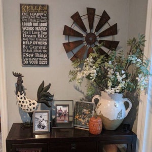 May include: A rustic farmhouse style decor vignette with a wooden sign with the text "Kelley Family Rules" and a list of rules, a metal windmill, a white ceramic vase with white flowers, a rooster figurine, and two framed photos on a dark brown wooden table.