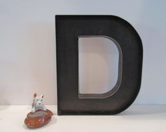 May include: A large, black metal letter D, standing upright on a white surface. The letter is made of thick metal and has a simple, industrial design.