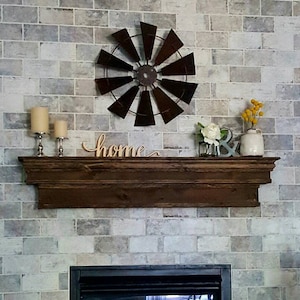 May include: A rustic wooden mantel shelf with a decorative metal windmill, candles, and a wooden sign that says "home". The shelf is mounted on a grey brick wall.