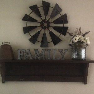 May include: A dark brown wooden shelf with a metal cowbell, a metal windmill, and the word "FAMILY" in metal letters. A silver metal vase with white flowers sits on the shelf.