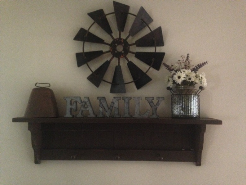 May include: A dark brown wooden shelf with a metal cowbell, a metal windmill, and the word "FAMILY" in metal letters. A silver metal vase with white flowers sits on the shelf.