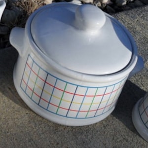 May include: White ceramic teapot, lidded casserole dish, and creamer. Each piece has a grid pattern in blue, red, yellow, and green. The teapot has a spout and handle. The casserole dish has two handles. The creamer has two handles.