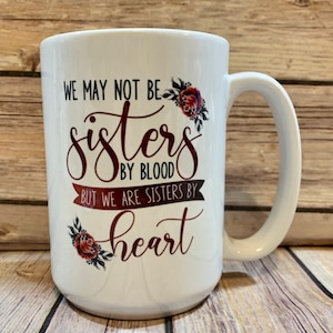 May include: White ceramic mug with a red and black floral design and the text "We may not be sisters by blood but we are sisters by heart".