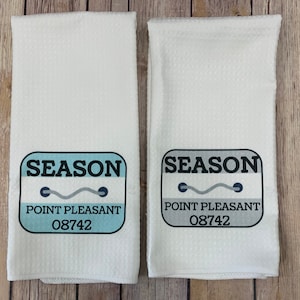 May include: Two white waffle-weave hand towels. Each towel features a rectangular design with the word "SEASON" above "POINT PLEASANT 08742". The design includes a wavy line and two dots. The text is in black, blue, and gray.