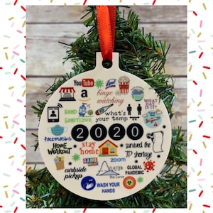 May include: A white round Christmas ornament with a 2020 design. The ornament features various images and text related to the year 2020, including "stay home", "zoom", "facemasks", "hand sanitizer", "global pandemic", and "wash your hands".