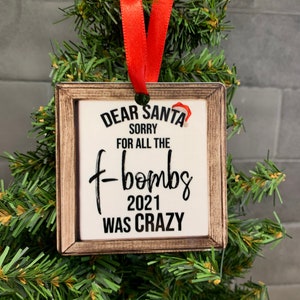 May include: A wooden Christmas ornament with a white background and the text "Dear Santa Sorry for all the f-bombs 2021 was crazy". The ornament has a red ribbon attached to the top.