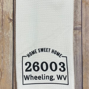 May include: White kitchen towel with black text that reads "Home Sweet Home 26003 Wheeling, WV".