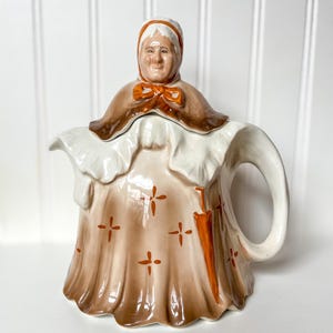 May include: A vintage ceramic teapot in the shape of a woman. The teapot features a brown shawl, a white bonnet, and a tan dress adorned with orange cross-shaped designs. An orange umbrella is painted on the side. The handle and spout are white.