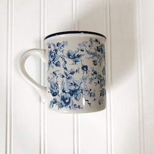 May include: A ceramic mug with a cream-colored base featuring a detailed blue floral pattern of roses and vines. The mug has a curved handle on the left side and a dark blue rim along the top edge, providing a clear view of the exterior design.