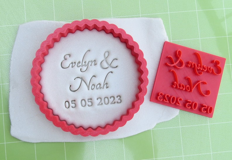 May include: Red plastic cookie cutter with a scalloped edge, featuring the text "Evelyn & Noah 05 05 2023". A separate red plastic stamp with the same text is also shown.