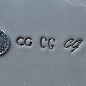 May include: Close-up of a gray surface with four impressions of the letters "CG" in different styles. The letters are stamped into the surface.