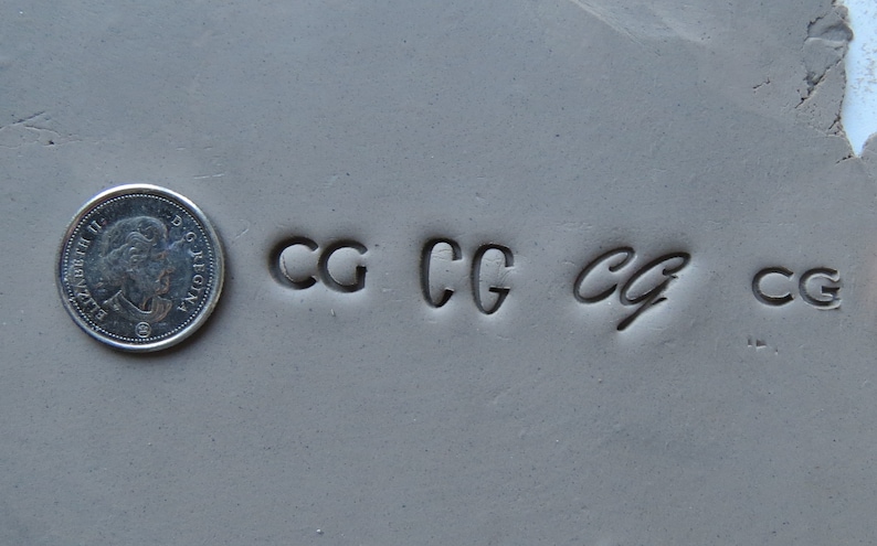 May include: Close-up of a gray surface with four impressions of the letters "CG" in different styles. The letters are stamped into the surface.