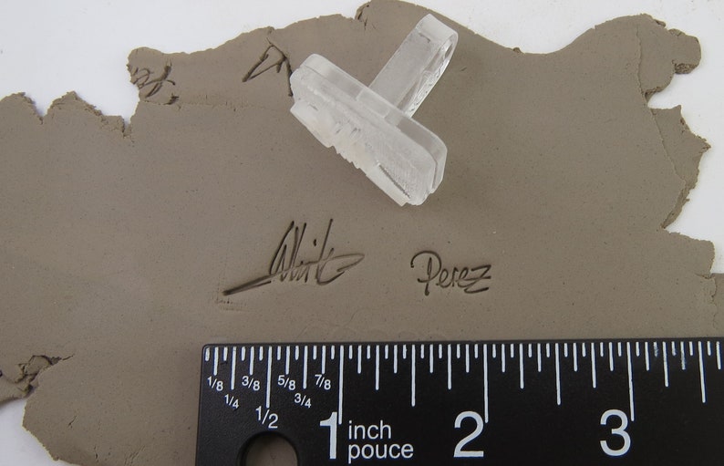 Pu&ograve; includere: Un timbro rettangolare trasparente con un design strutturato poggia su un pezzo di argilla. L'argilla &egrave; contrassegnata dalle firme "Whit" e "Perez". Un righello mostra le misure in pollici.