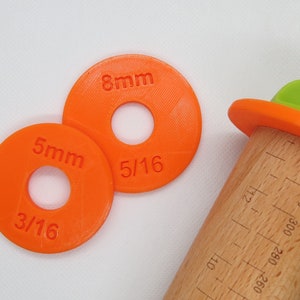 May include: Two orange plastic rings with different sized holes. The rings are labelled with measurements in millimetres and fractions of an inch. The rings are used for rolling out dough to a specific thickness.