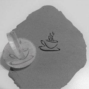 May include: A clear plastic stamp with a design of a coffee cup with steam rising from it. The stamp is being used to create an impression in a grey clay surface.