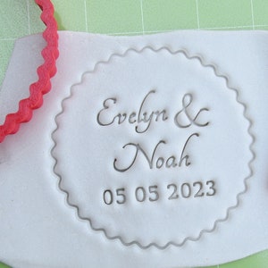 May include: A white fondant circle with a scalloped edge, imprinted with the text "Evelyn & Noah 05 05 2023". A red rubber stamp with the same text is also shown.