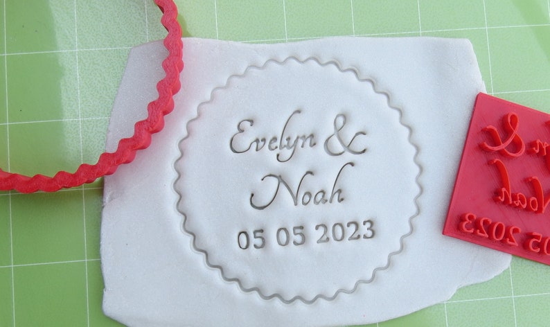 May include: A white fondant circle with a scalloped edge, imprinted with the text "Evelyn & Noah 05 05 2023". A red rubber stamp with the same text is also shown.