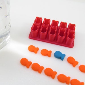 May include: A red plastic mould with 12 fish-shaped cavities, a glass of water, and orange and blue fish-shaped sweets.