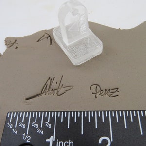 Pu&ograve; includere: Uno strumento di timbratura in plastica trasparente &egrave; appoggiato su una superficie di argilla grigia. L'argilla presenta la firma dell'artista e il nome "Perez" incisi. Un righello mostra le misure in pollici.