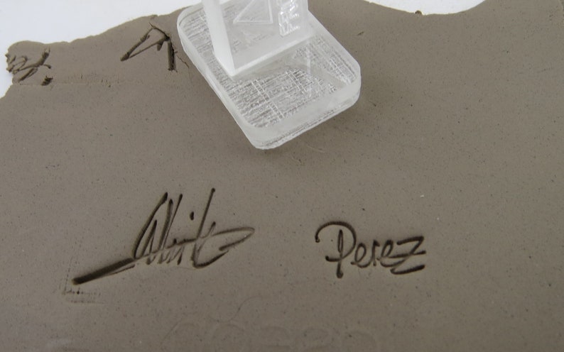 Pu&ograve; includere: Un pezzo di ceramica grigio chiaro, di forma irregolare, con una struttura geometrica trasparente sulla parte superiore. Il pezzo &egrave; firmato con i nomi "Whitt" e "Perez" in grafia nera.