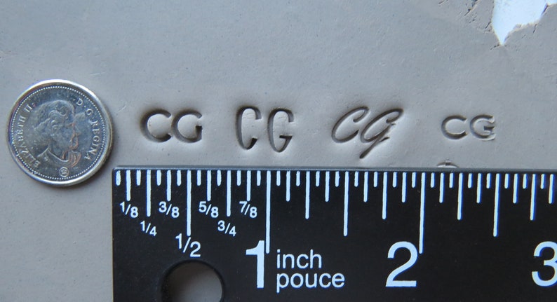 May include: Close-up of a ruler measuring the size of four different "CG" stamps pressed into clay. The ruler is marked in inches and shows the stamps are about 1/2 inch wide.