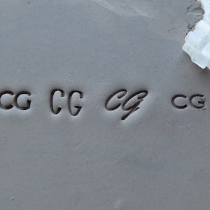 May include: A close-up of a gray clay surface with four impressions of the letters "CG" made with a clear plastic stamp. The stamp is labeled "FRONT" and is visible in the upper right corner of the image. A penny is visible in the lower left corner of the image.