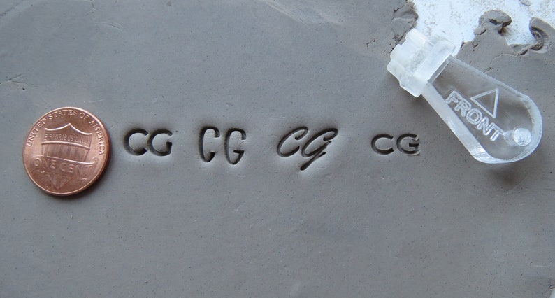 May include: A close-up of a gray clay surface with four impressions of the letters "CG" made with a clear plastic stamp. The stamp is labeled "FRONT" and is visible in the upper right corner of the image. A penny is visible in the lower left corner of the image.