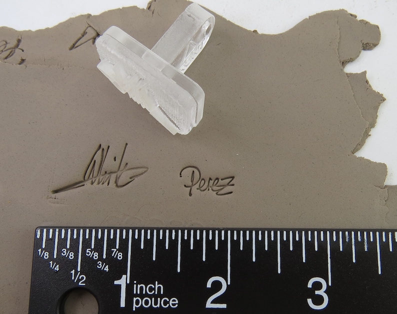 Pu&ograve; includere: Un timbro rettangolare trasparente con un design dettagliato poggia su una superficie di argilla grigia. Il timbro &egrave; posizionato vicino alla firma "Witte Perez" e un righello che mostra pollici. L'argilla ha una consistenza ruvida e irregolare.