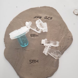 May include: A close-up of a light brown clay surface with three clear plastic stamps and one blue plastic stamp. The stamps are labeled "APR CLB", "HBS", and "FRONT".