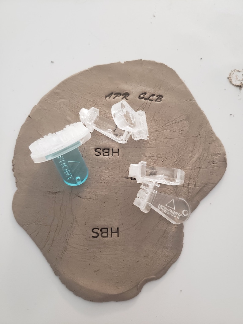 May include: A close-up of a light brown clay surface with three clear plastic stamps and one blue plastic stamp. The stamps are labeled "APR CLB", "HBS", and "FRONT".