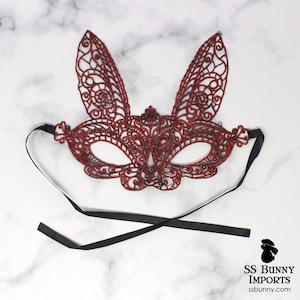 May include: Red lace bunny mask with black ribbon ties. The mask has a delicate, intricate design.