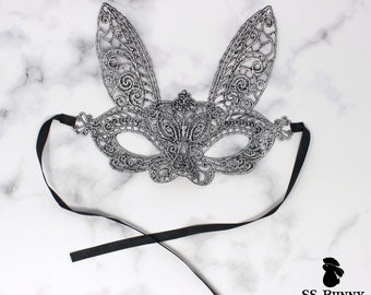 May include: A silver lace bunny mask with black ribbon ties. The mask has a delicate, intricate design.